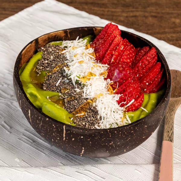 A vibrant green smoothie bowl in a coconut shell, topped with strawberries, chia seeds, and shredded coconut.
