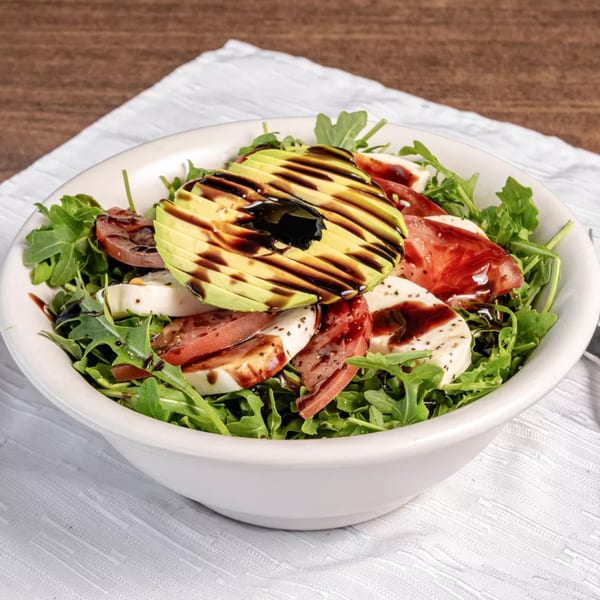 A healthy salad with arugula, sliced avocado, tomatoes, mozzarella, and balsamic glaze in a white bowl.