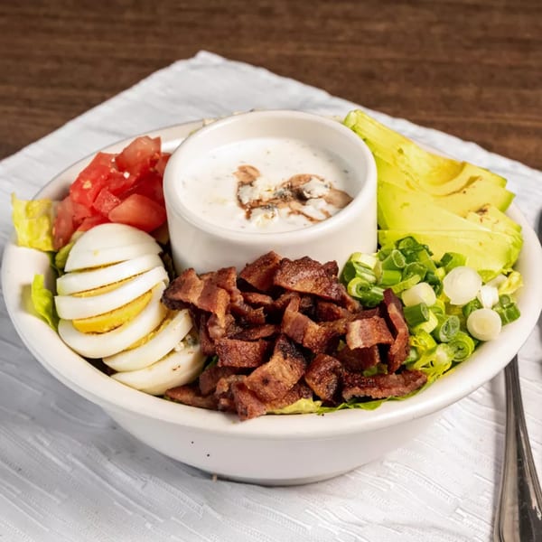 A healthy Cobb salad with bacon, avocado, tomatoes, hard-boiled eggs, and dressing in a white bowl.