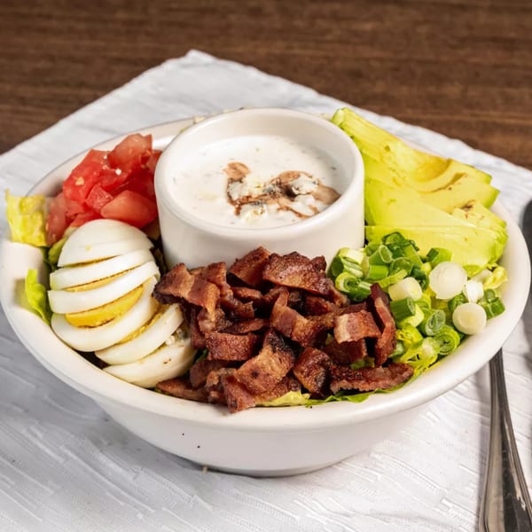 A healthy Cobb salad with bacon, avocado, tomatoes, eggs, and dressing in a white bowl.
